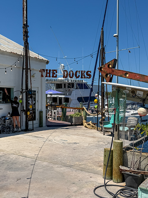 Entrance to The Docks restaurant at a marina with boats in the background and string lights overhead in a sunny scene.