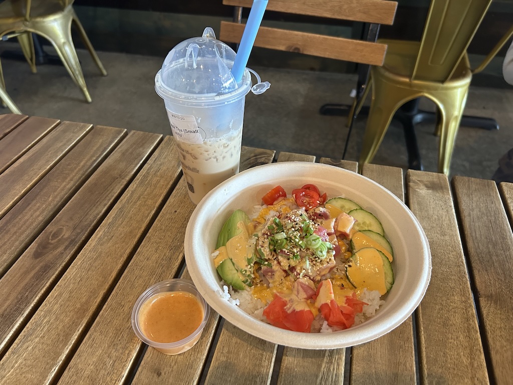 Poke bowl with rice, tuna, mango, cucumber, tomato, sesame seeds and green onions on a wooden table; side sauce cup and iced coffee drink nearby.